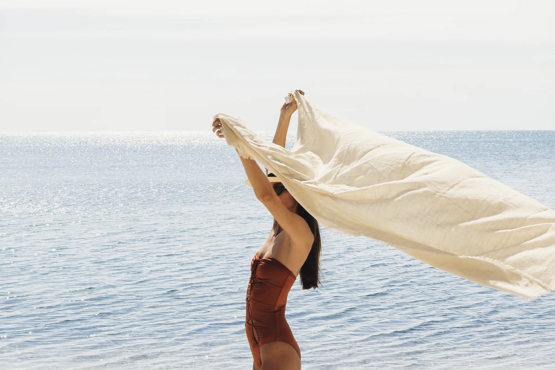 Person in rust swimsuit holds billowing fabric at ocean’s edge under clear sky.