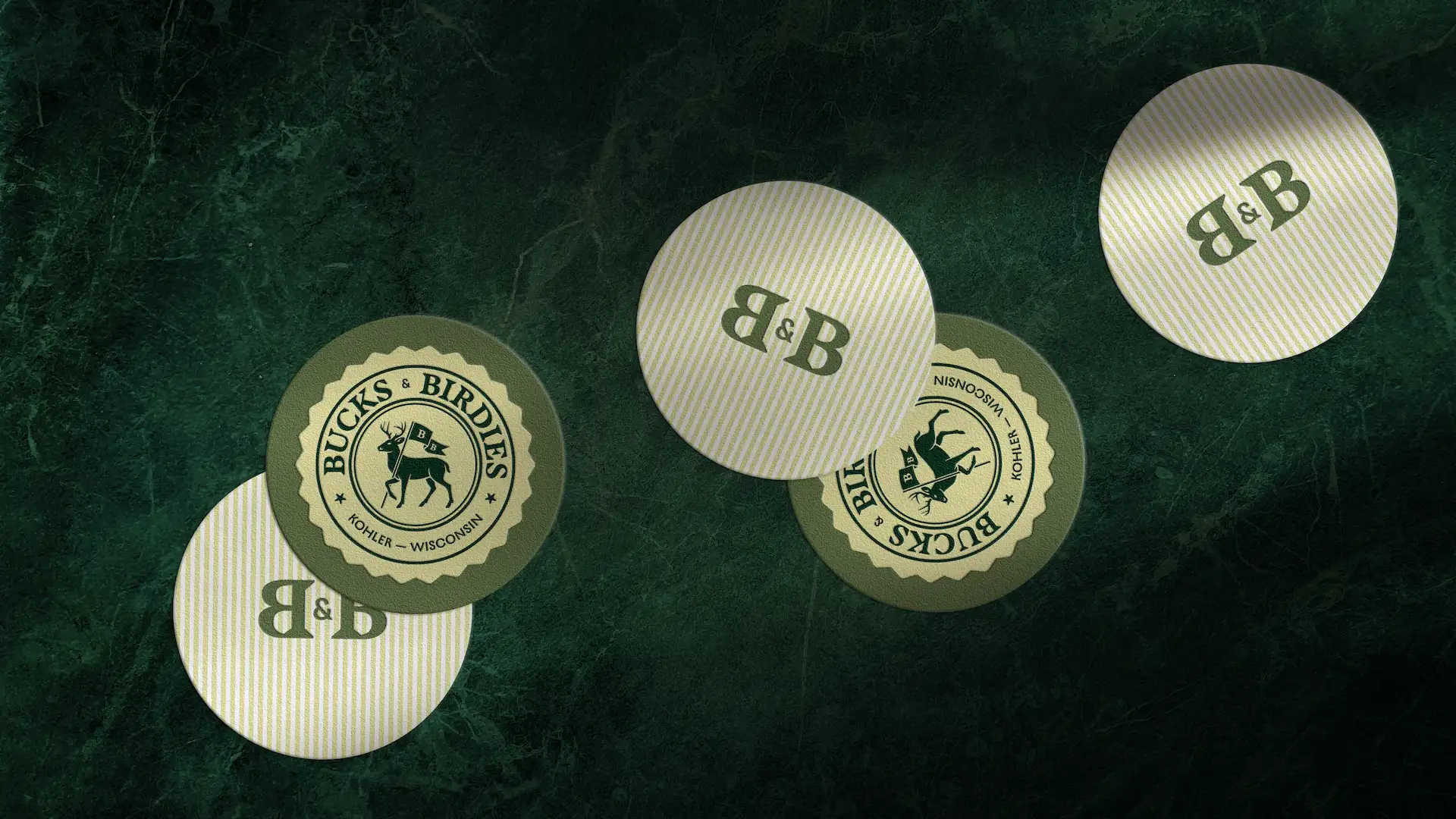 Five round coasters on green marble: three striped with “B & B” text, two with Bucks & Birdies deer logo.