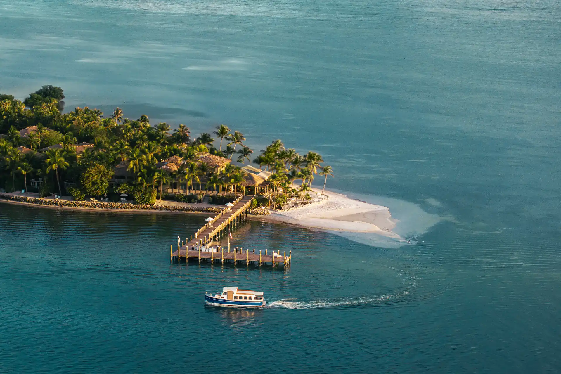 Aerial view of a private tropical island with palm trees, sandy shore, wooden pier, and a boat approaching.