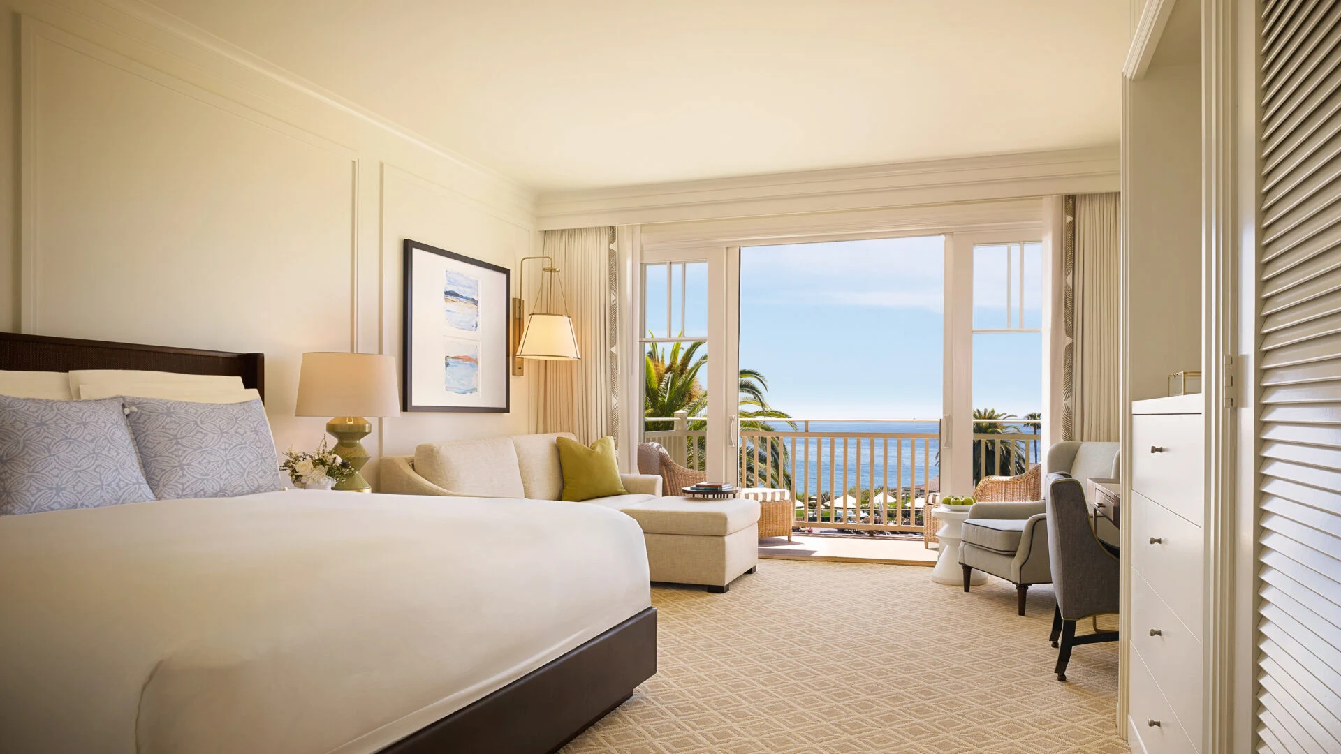 Bright hotel bedroom with balcony ocean view, palm trees outside, king bed, sofa seating, and soft neutral decor.