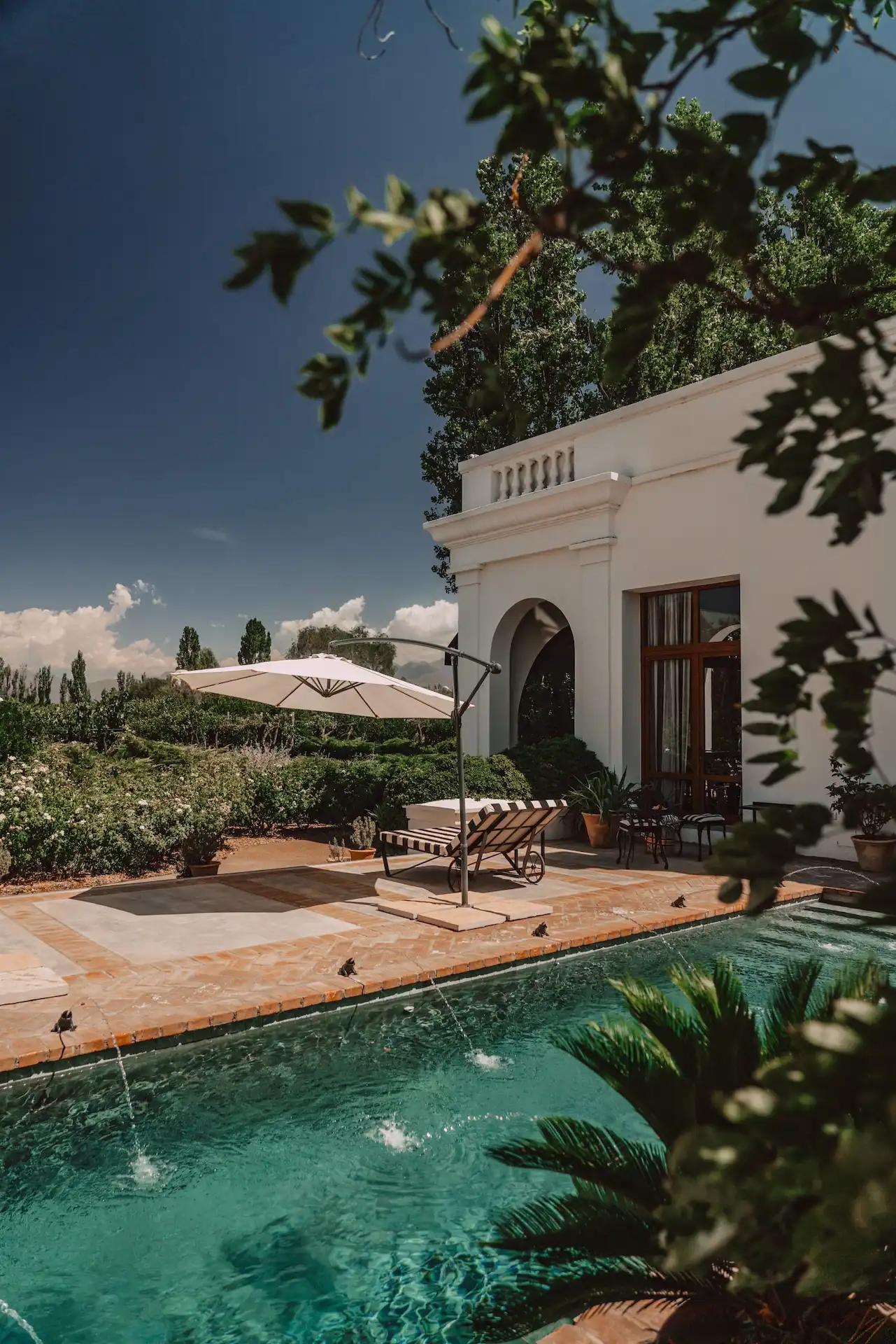 Websites by Spherical 4 Pool with water jets, striped lounge chair, umbrella, lush greenery, and white building with columns.