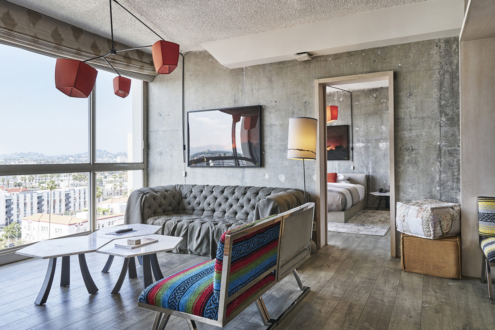 Websites by Spherical 2 Modern hotel suite with concrete walls, city view windows, sofa seating area, and bedroom visible through an open doorway.