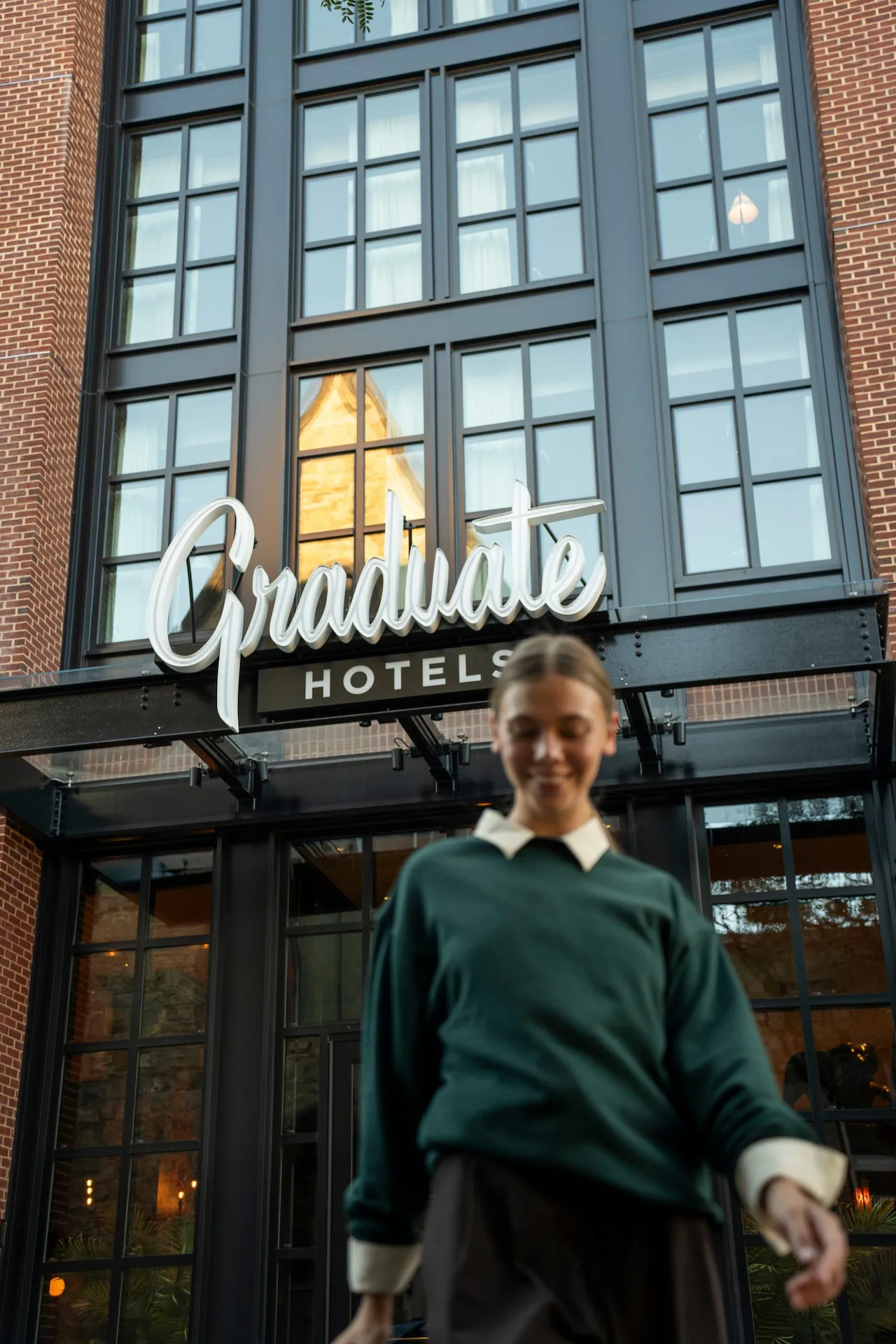 Smiling girl in green sweater and white shirt walking in front of Graduate Hotels facade.