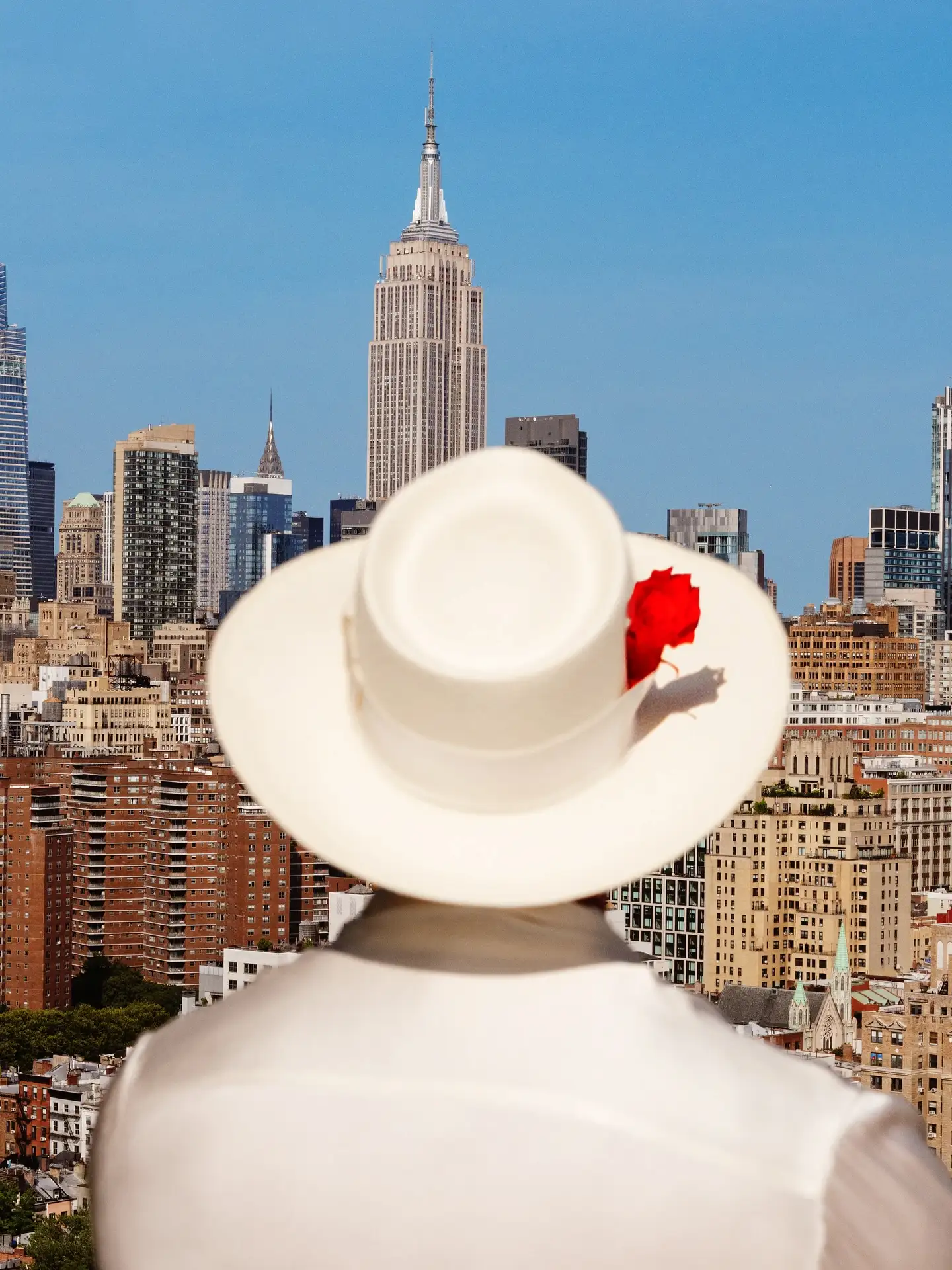 Person in white hat with red flower facing Empire State Building under clear blue sky