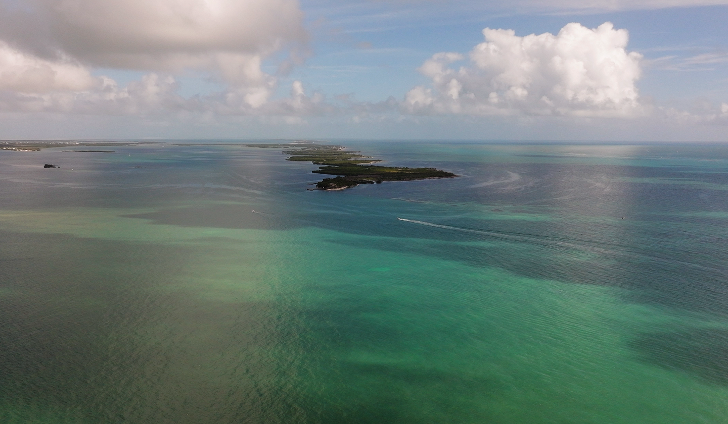 Aerial view of turquoise ocean with scattered islands, gentle waves, and clouds stretching toward the horizon.
