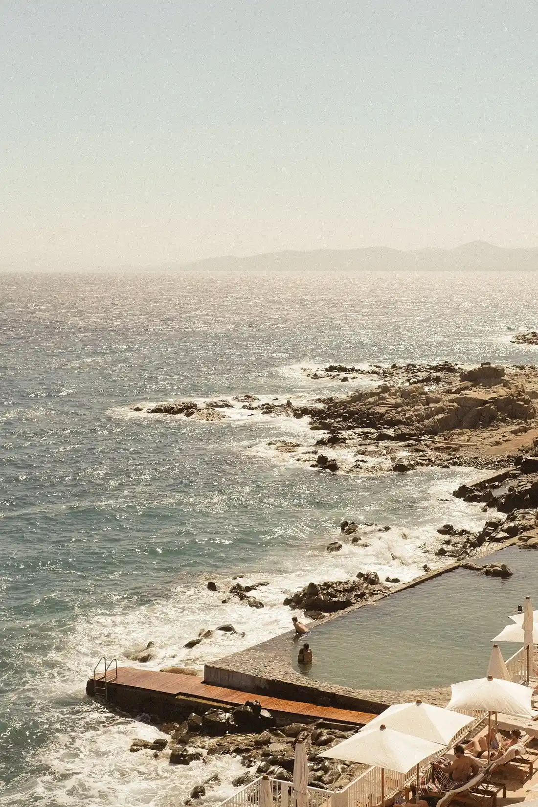 Infinity pool at Baumier overlooking rocky coastline with white umbrellas, lounge chairs, and ocean horizon