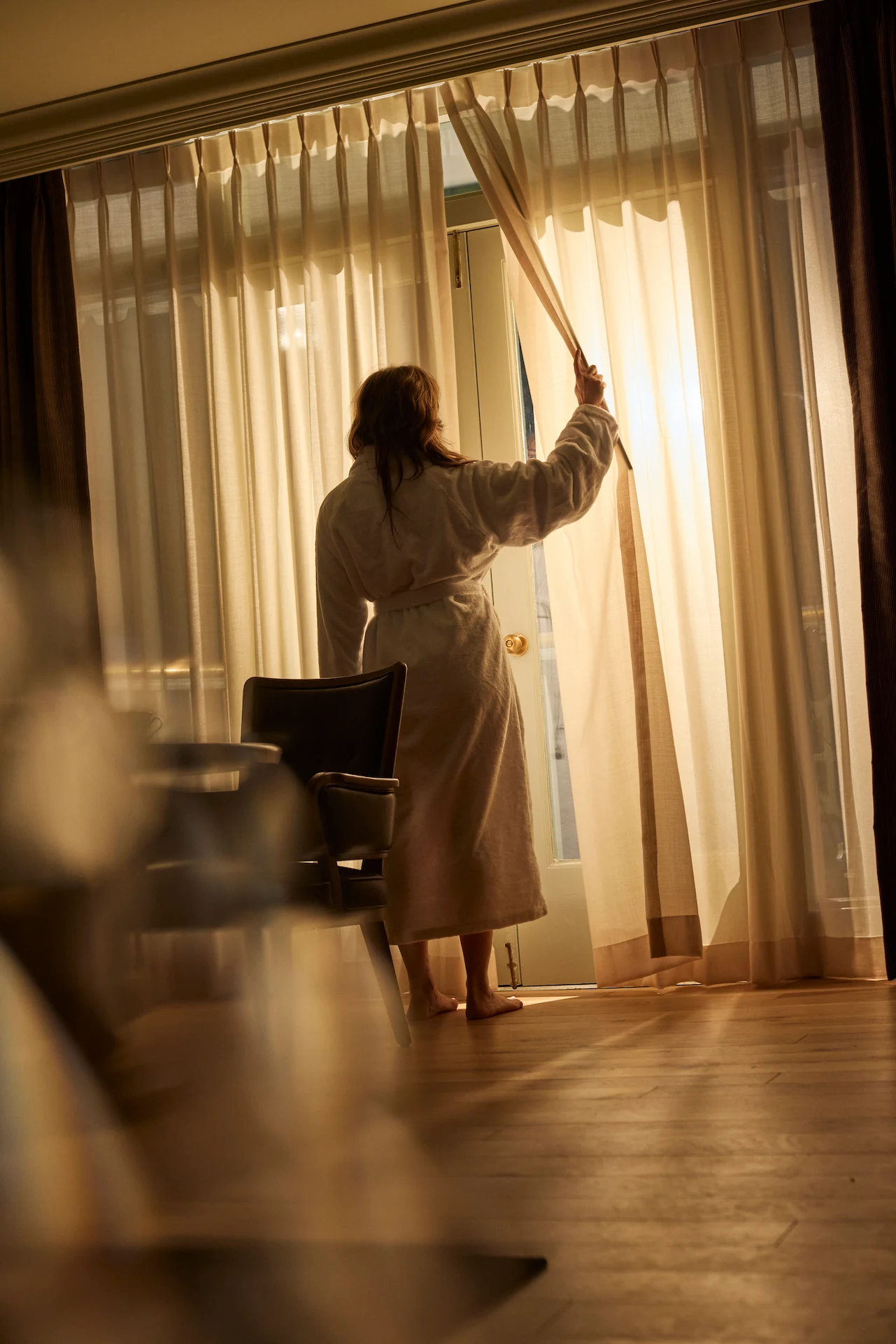 Guest in bathrobe opening curtains at The Bellevue Hotel, morning light streaming through window onto wooden floor.
