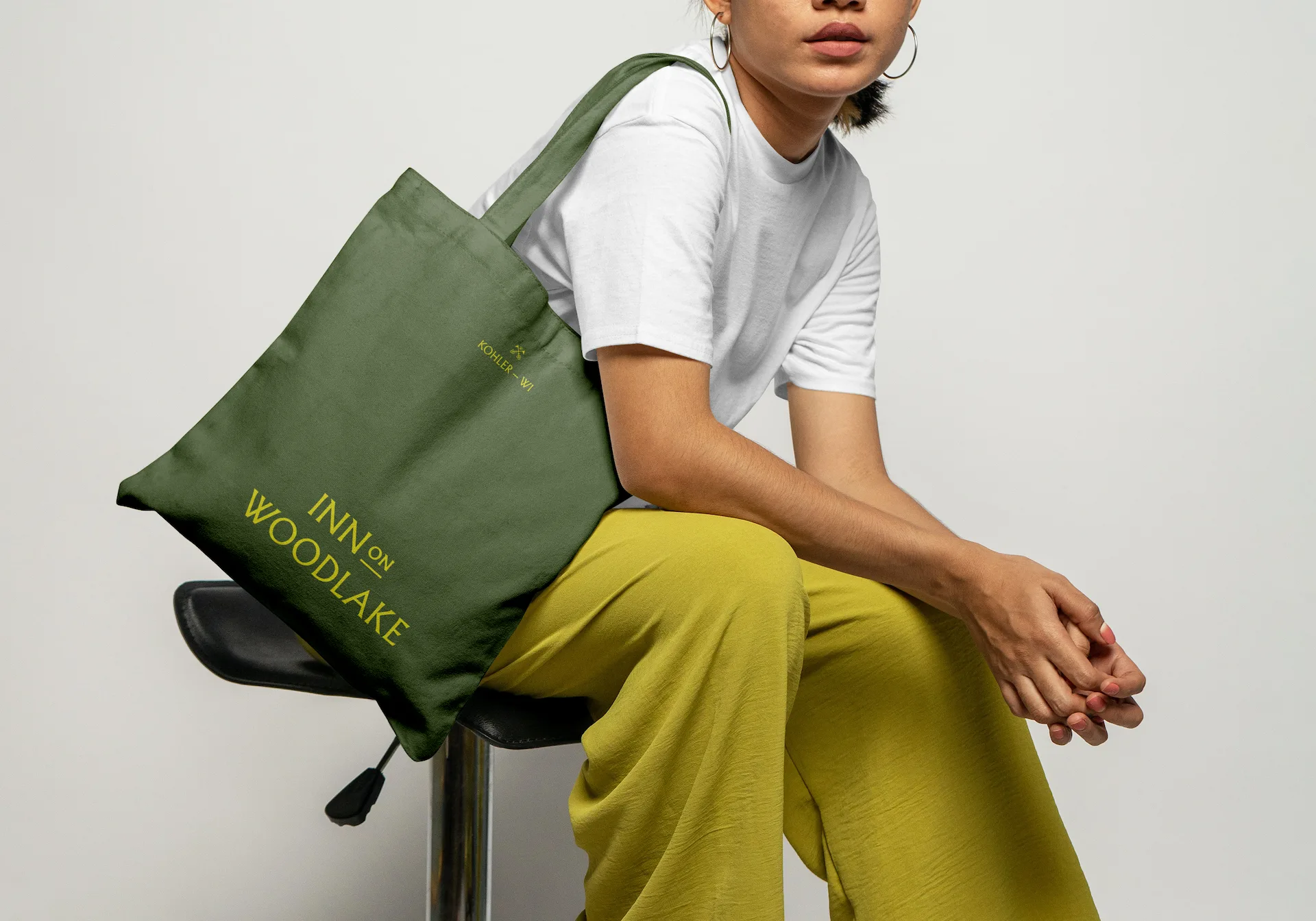Person seated on stool wearing white shirt & yellow-green pants, holding green tote bag labeled “Inn on Woodlake, Kohler WI."